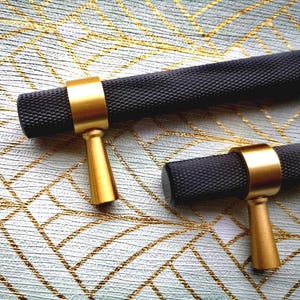 May include: Close-up of two black cabinet pulls with a textured surface and gold-colored accents. The pulls have a cylindrical shape with a gold ring and a gold handle. The hardware is set against a patterned background.
