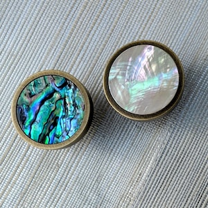 May include: Two circular cabinet knobs with brass-toned rims. One displays a vibrant, iridescent abalone shell design in blues and greens. The other has a pearlescent white and iridescent surface.