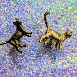 May include: Two cat figurines. One is bronze-colored, posed with front paws extended. The other is gold-colored, with its tail raised. Both are detailed with textured fur. They are set against a sparkly, iridescent background.