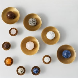 Puede incluir: Un surtido de cuencos y esferas decorativas. Los cuencos son de color dorado, y las esferas varían en color y textura, incluyendo marrón, moteado, rosa, blanco, naranja y azul. La disposición se muestra sobre un fondo blanco.