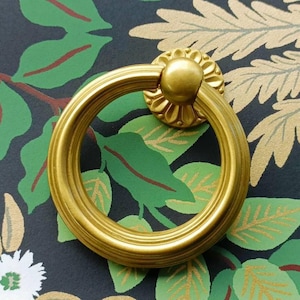 以下が含まれることがあります： 金色の円形引き出し取っ手、花のバックプレート付き。 引き手は、緑の葉と金のフローラルパターンを背景に配置され、装飾的なハードウェア要素を作成しています。