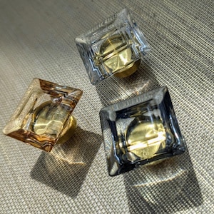 May include: Three square glass cabinet knobs with gold-coloured bases. The knobs have a faceted design, with one in amber, one in clear, and one in a smoky grey colour. The knobs are arranged on a textured surface.