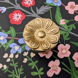 Könnte beinhalten: Ein goldfarbener, blütenförmiger Schrankknopf. Der Knopf ist auf einem schwarzen Hintergrund mit einem Blumenmuster aus roten, rosa, blauen und weißen Blumen und grünen Blättern zentriert. Der Knopf hat eine detaillierte, strukturierte Oberfläche.