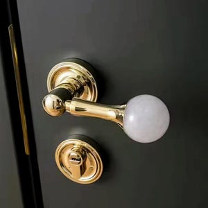May include: A gold door handle with a white, round knob. The handle has a keyhole and a decorative design.