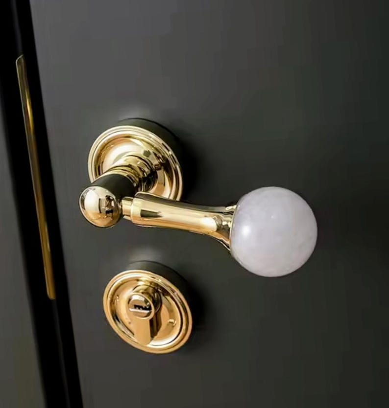 May include: A gold door handle with a white, round knob. The handle has a keyhole and a decorative design.