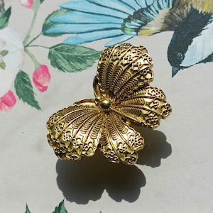 Puede incluir: Un broche de mariposa dorado y ornamentado con detalles intrincados. El broche tiene una esfera central y alas simétricas en forma de abanico. El fondo presenta un diseño floral y de aves.