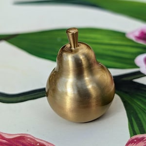 May include: A small, golden-coloured pear-shaped object with a stem. The pear is made of metal and has a smooth, polished surface. The pear is sitting on a floral patterned surface.