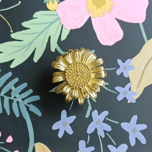 May include: A gold-colored metal daisy flower-shaped decorative object. The daisy has detailed petals and a textured center. The object is set against a dark background with floral and leaf patterns in pink, blue, and yellow.