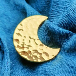 May include: A gold-coloured crescent moon-shaped brooch with a hammered texture. The brooch is set against a backdrop of blue fabric, creating a contrast in colour and texture. The moon shape is a common symbol.