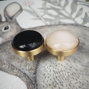 May include: Two cabinet knobs with a black and a cream-coloured dome. Both knobs have a gold-coloured base and stem. The knobs are likely for furniture or home decor. The knobs are likely for furniture or home decor.
