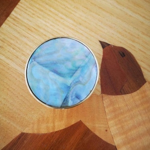 May include: A circular object with a silver rim and a blue and white marbled pattern. It sits on a wooden surface with a bird inlay. The bird is crafted from various shades of brown wood.