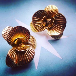May include: Two gold-colored flower-shaped rings. The rings have detailed petal designs and are set against a blue background with a white star-like shape. The rings appear to be made of metal.