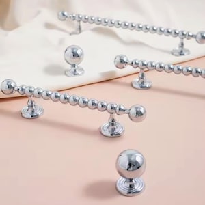 Puede incluir: Una colección de herrajes plateados brillantes para gabinetes, que incluyen varios tiradores de barra con cuentas y perillas redondas. El hardware se muestra sobre una superficie rosa claro con un paño blanco de fondo. Los tiradores tienen una serie de pequeñas cuentas redondas.