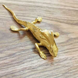 Puede incluir: Una figura de lagarto dorado con textura detallada, sobre una superficie de madera. El lagarto tiene cuatro patas, una cola larga y un cuerpo texturizado. Es un artículo decorativo.