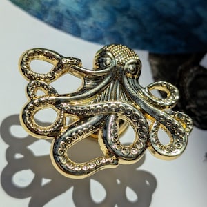 May include: A gold-colored octopus-shaped decorative object. The octopus has detailed tentacles and a textured head. The object is casting a shadow on a white surface. The overall design is intricate and detailed.