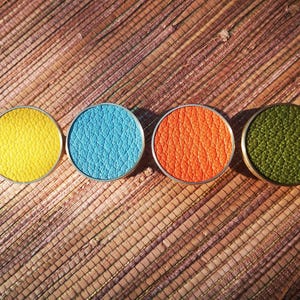 May include: Four round, colorful objects with textured surfaces and silver rims are arranged in a row. The colors are yellow, blue, orange, and green. The objects are set against a woven, brown background.