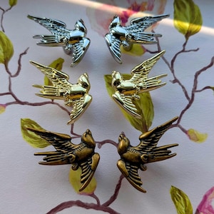 May include: Six metal bird-shaped decorative accents in silver, gold, and bronze tones. The birds are arranged in two rows of three, with wings outstretched. The background features a floral pattern with pink and green hues.