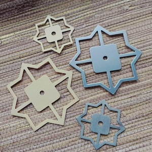 以下が含まれることがあります： 4つの金属製の星型の装飾品。2つは金色、2つは銀色です。それぞれ正方形の中心に小さな穴があり、スターバーストのデザインで囲まれています。それらは、テクスチャ加工された茶色の表面に配置されています。