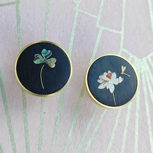 Puede incluir: Dos tiradores redondos para armarios con fondo azul oscuro y borde dorado. Un tirador presenta un diseño de trébol con detalles iridiscentes, y el otro tiene una flor blanca y una pequeña libélula. Estos tiradores decorativos añaden elegancia.