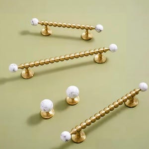 May include: A selection of gold-coloured cabinet hardware, including pulls and knobs. The pulls feature a series of small, round beads and are accented with white marble-patterned end pieces. The knobs have a similar marble design.