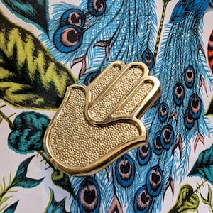 Peut inclure: Un objet décoratif doré en forme de main avec une surface texturée. L'objet est posé sur un fond de motif coloré de plumes de paon dans des tons de bleu, vert et blanc.