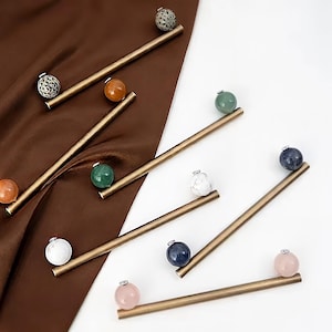 May include: A collection of six linear metal cabinet pulls in a brushed brass finish, each featuring a single spherical gemstone knob. The stones include spotted, orange, green, white, blue, and pale pink varieties, attached to the ends.