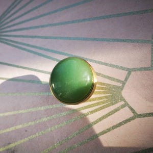 May include: A circular, green gemstone ring with a gold-coloured band. The gemstone has a smooth, polished surface and a gradient of green shades. The ring is set against a background with a geometric pattern.