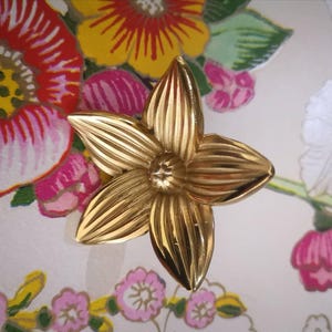 May include: A gold-colored, five-petal flower-shaped decorative object. The petals have a ribbed texture, and the center has a small, star-like design. The background features a floral pattern with red, pink, yellow, and white flowers.