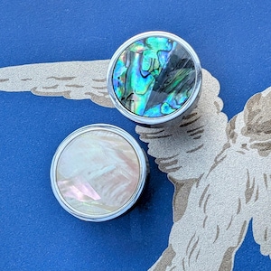 May include: Two round buttons with silver rims. One button has iridescent abalone shell, showing blue, green, and black hues. The other button has a pearlescent white shell. The buttons are on a blue surface with a silver wing design.