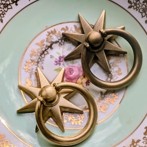 May include: Two antique brass starburst drawer pulls with ring handles. The pulls are set on a vintage plate with a light green centre, gold trim, and floral accents. The starburst design adds a touch of elegance.
