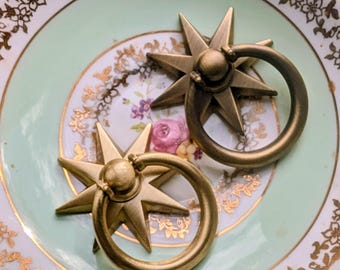 Könnte beinhalten: Zwei antike Schubladengriffe aus Messing in Sternform mit Ringgriffen. Die Griffe liegen auf einem Vintage-Teller mit hellgrüner Mitte, goldenem Rand und floralen Akzenten. Das Stern-Design verleiht einen Hauch von Eleganz.