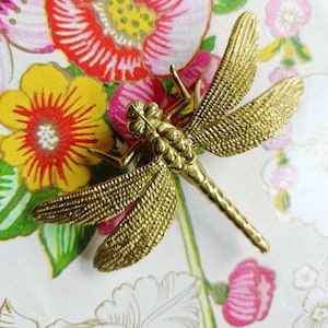 May include: A gold-toned dragonfly brooch with intricate wing and body detailing. The dragonfly is positioned on a background of colourful floral designs in red, yellow, and pink, offering a striking contrast.
