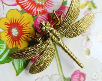 Puede incluir: Un broche de libélula dorada con alas y cuerpo texturizados. La libélula se encuentra sobre un fondo de coloridos diseños florales en rojo, amarillo y rosa, creando un contraste vibrante.
