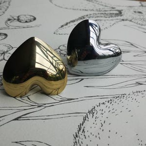 May include: Two heart-shaped decorative objects. One is gold-toned, the other is silver-toned. Both have a smooth, reflective surface. The objects are placed on a white surface with a black and white floral pattern.