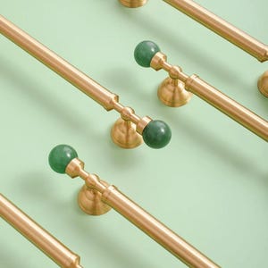 May include: Close-up of several gold-toned cabinet pulls with a green, spherical accent. The pulls are mounted on a light green surface. The hardware has a classic design, with a long bar and decorative base.