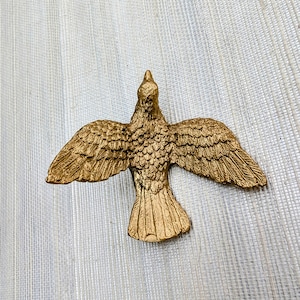 May include: A gold-colored bird sculpture with outstretched wings. The bird is detailed with textured feathers and a pointed beak. The sculpture is likely made of metal and is displayed against a neutral background.