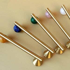 May include: A collection of brass cabinet pulls with colorful gemstone accents. Each pull features a cylindrical brass bar and a round base. The gemstones include yellow, blue, green, pink, and white, adding a touch of elegance to any furniture.