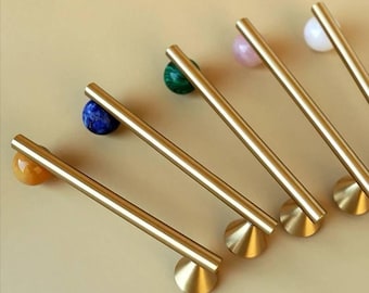 May include: Five gold-toned metal cabinet pulls arranged in a diagonal row. Each pull features a long, cylindrical bar attached to a conical base at one end and a small, spherical gemstone accent at the other, including yellow, blue, green, pink, and white.