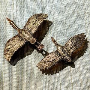 May include: Two bronze-coloured crane figurines in flight. The birds have outstretched wings, detailed feathers, and long necks. The figurines are set against a textured, neutral-toned background, creating a contrast that highlights their form.