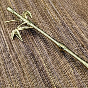 May include: A gold-colored bamboo-shaped decorative object. The object has a long, slender body with a series of nodes and leaf-like details. The bamboo-shaped object is made of a shiny, metallic material and is set against a textured, brown background.