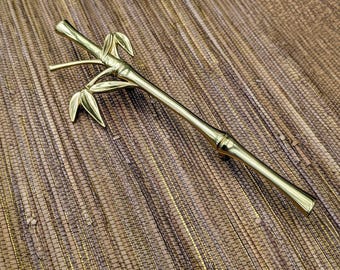 May include: A gold-colored bamboo-shaped decorative object. The object has a long, slender body with a series of nodes and leaf-like details. The bamboo-shaped object is made of a shiny, metallic material and is set against a textured, brown background.
