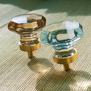 May include: Two faceted glass cabinet knobs with brass bases. One knob has a warm amber hue, while the other is a cool, clear blue. The knobs are designed for furniture and add a touch of elegance.
