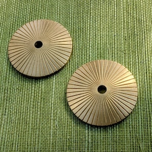 以下が含まれることがあります： サンバーストデザインの真鍮色の丸いコースター2つ。各コースターには中央の穴と放射状の溝があります。コースターは、質感のある緑色の背景に表示されています。
