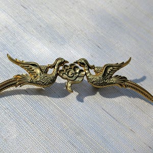 May include: A gold-coloured brooch depicting two stylised birds facing each other, with outstretched wings and long tail feathers. The birds are connected by a floral design. The brooch is made of a shiny, metallic material.