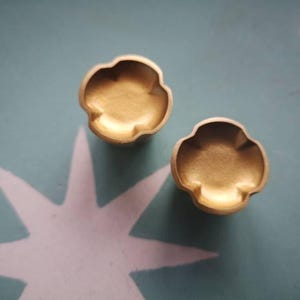 Könnte beinhalten: Zwei kleine, goldfarbene, blütenförmige Objekte. Die Objekte haben ein abgerundetes, blütenblattartiges Design und eine leicht konkave Mitte. Sie befinden sich auf einem türkisblauen Hintergrund mit einem weißen Sternmuster.