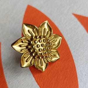 May include: A gold-colored sunflower-shaped brooch. The brooch has detailed petals and a textured center, resembling a real sunflower. The brooch is a jewelry item.