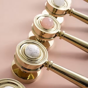 May include: Five gold door knobs with different coloured stone inlays. The knobs are arranged in a row on a pink background. The stones are white, pink, and a light pink.