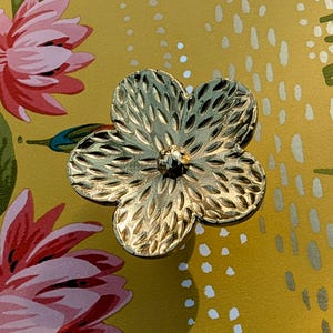 May include: A gold-coloured, flower-shaped decorative knob with a textured surface. The knob has a small, dark centre and is set against a yellow background with a floral pattern featuring pink and green flowers.