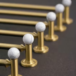 Puede incluir: Un conjunto de tiradores dorados para armarios con pomos esféricos de mármol blanco y gris. Los tiradores están dispuestos en fila, mostrando su diseño decorativo y su potencial para herrajes de cocina o baño.