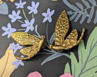 Puede incluir: Dos broches decorativos dorados con forma de libélula. Los broches tienen patrones de alas detallados y están colocados sobre un fondo oscuro con diseños florales y de hojas en azul, verde y rosa.
